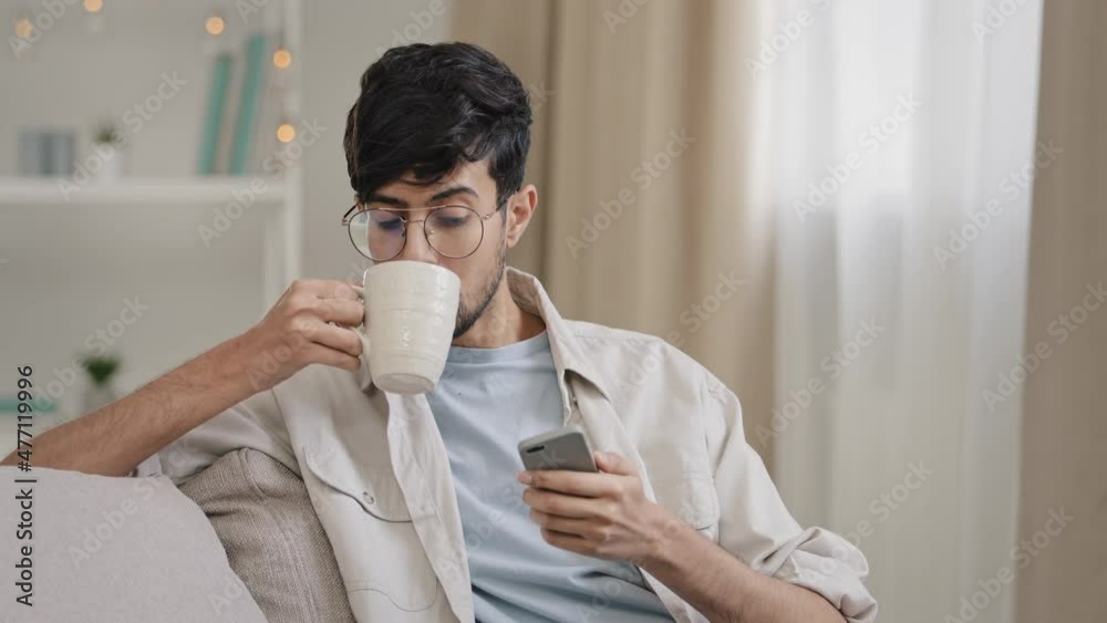 Young relaxed guy indian arabic bearded handsome man sitting at home on couch drinking tea coffee from cup looking at phone smiling watching funny video online chatting browsing with smartphone Young relaxed guy indian arabic bearded handsome man sitting at home on couch drinking tea coffee from cup looking at phone smiling watching funny video online chatting browsing with smartphone