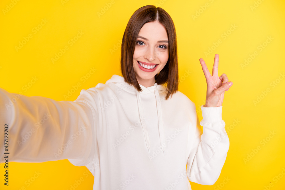 Photo of positive friendly lady take selfie show v-sign wear white hoodie isolated yellow color background Photo of positive friendly lady take selfie show v-sign wear white hoodie isolated yellow color background