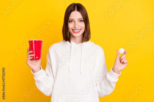 Photo of cheerful lady hold plastic cup tennis ball beer pong wear white hoodie isolated yellow color background photo