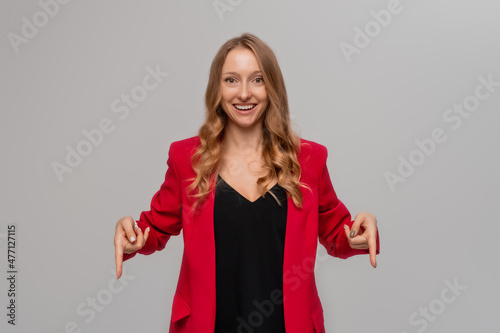 Smiling excited blonde young woman pointing fingers to the down, looking happy, showing to the advertisement, demonstrating promo offer, stands in red blazer against gray background