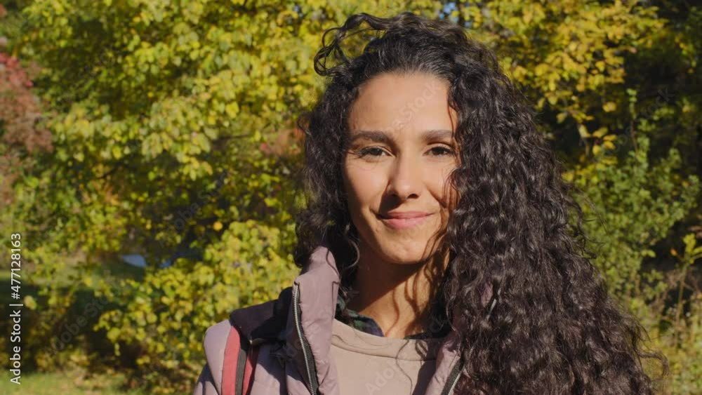Pretty millennial hispanic woman looking at camera with white-toothed smile standing at green forest trees background. Happy girl student fixes curly long hair, young beautiful lady closeup portrait