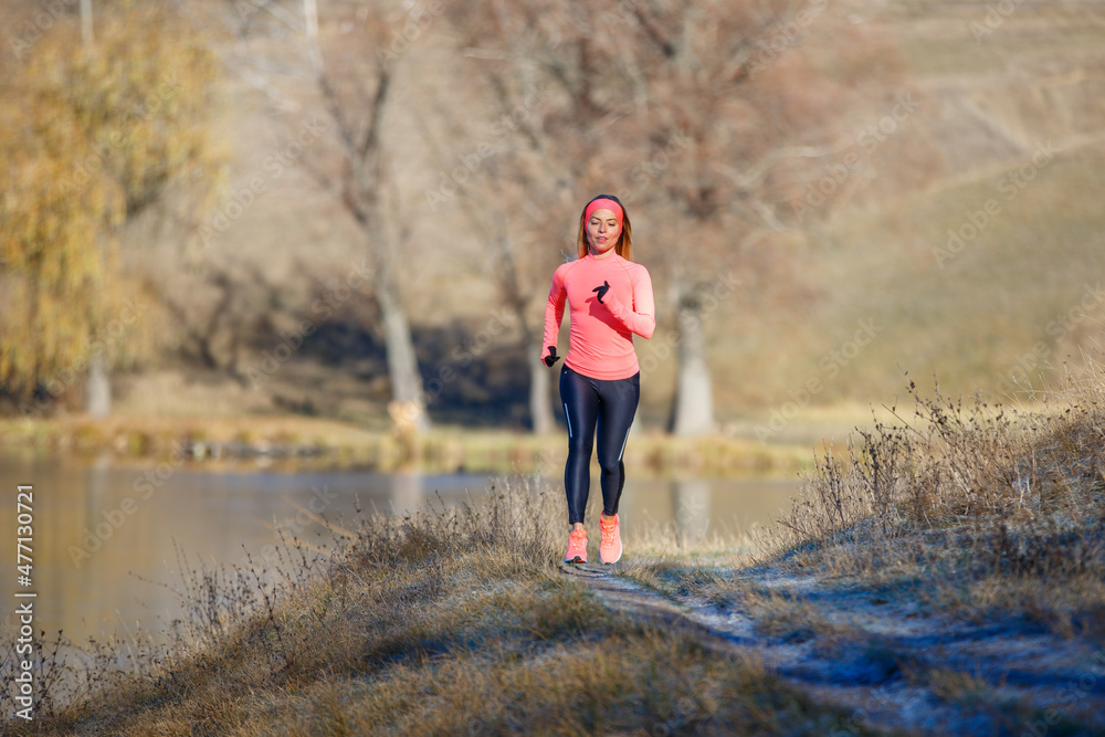 Fitness girl jogging in park near pond in the frosty morning. Running woman training near the lake in cold weather Fitness girl jogging in park near pond in the frosty morning. Running woman training near the lake in cold weather