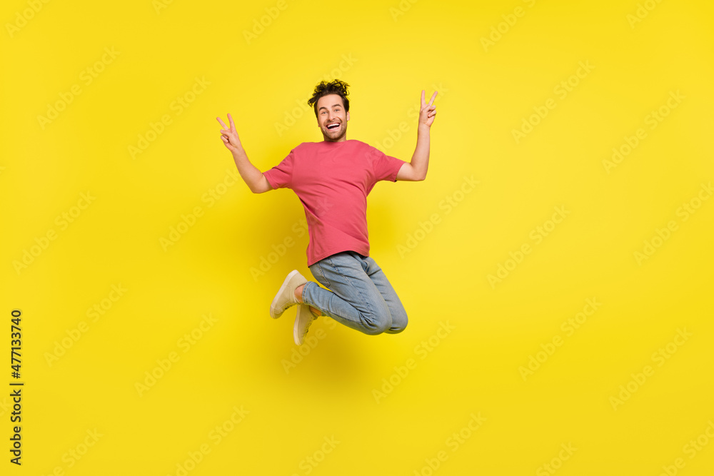 Full size photo of impressed brunet young guy jump show v-sign wear t-shirt jeans sneakers isolated on yellow background