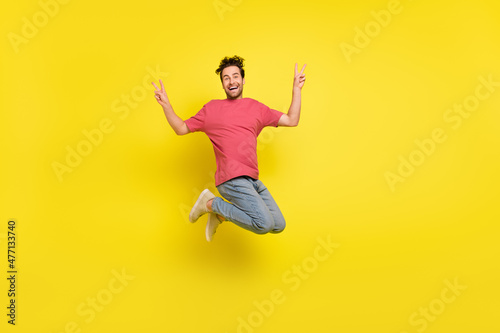 Full size photo of impressed brunet young guy jump show v-sign wear t-shirt jeans sneakers isolated on yellow background © deagreez