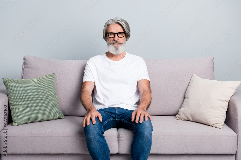 Portrait of attractive serious gray-haired man bachelor sitting on sofa alone isolated over grey pastel color background Portrait of attractive serious gray-haired man bachelor sitting on sofa alone isolated over grey pastel color background
