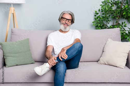 Photo of cute funny retired man wear white t-shirt glasses relaxing sitting coach smiling indoors isolated grey color background