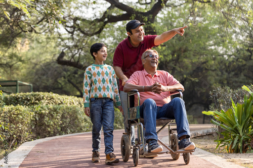 Man pushing old father on wheelchair while admiring view with son at park
