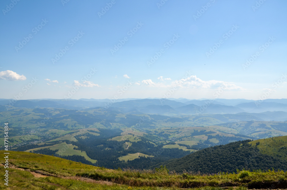 Fototapeta premium Beautiful rural landscape from the mountain range to the valley with green hills, grassy meadow, forest and village on a sunny summer day. Carpathians, Ukraine