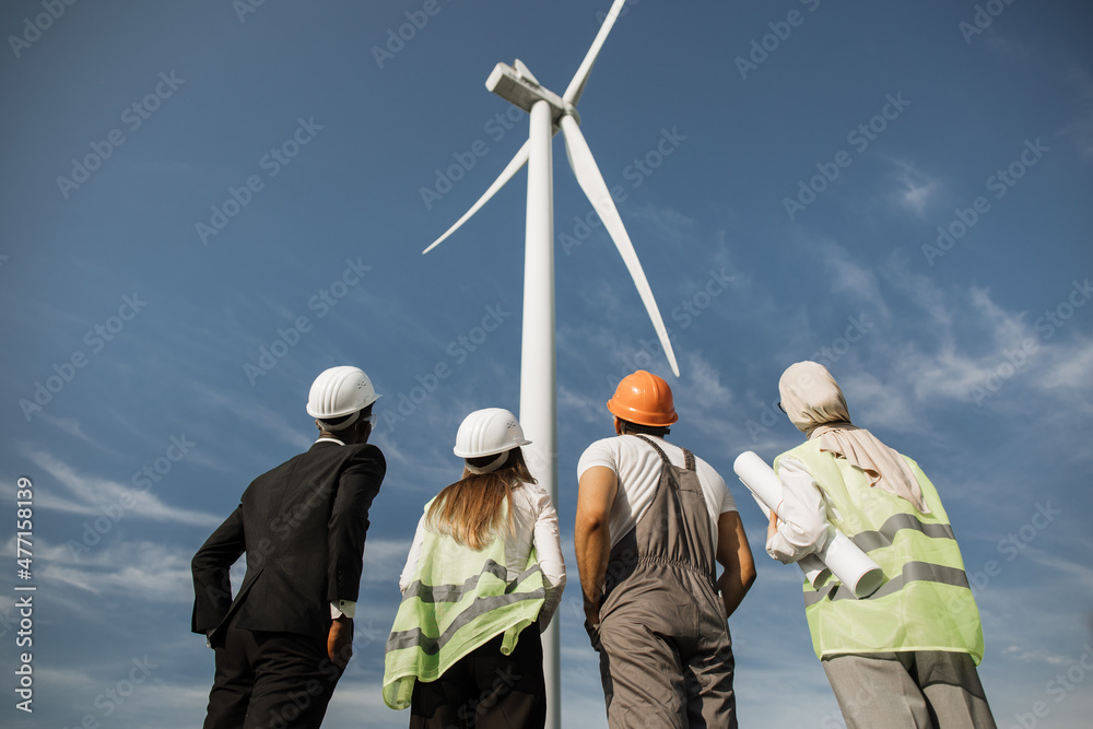 Back view of men and women in uniform and safety helmets looking on huge wind turbine while standing at rural area. Four multicultural partners discussing production of clean energy. Back view of men and women in uniform and safety helmets looking on huge wind turbine while standing at rural area. Four multicultural partners discussing production of clean energy.