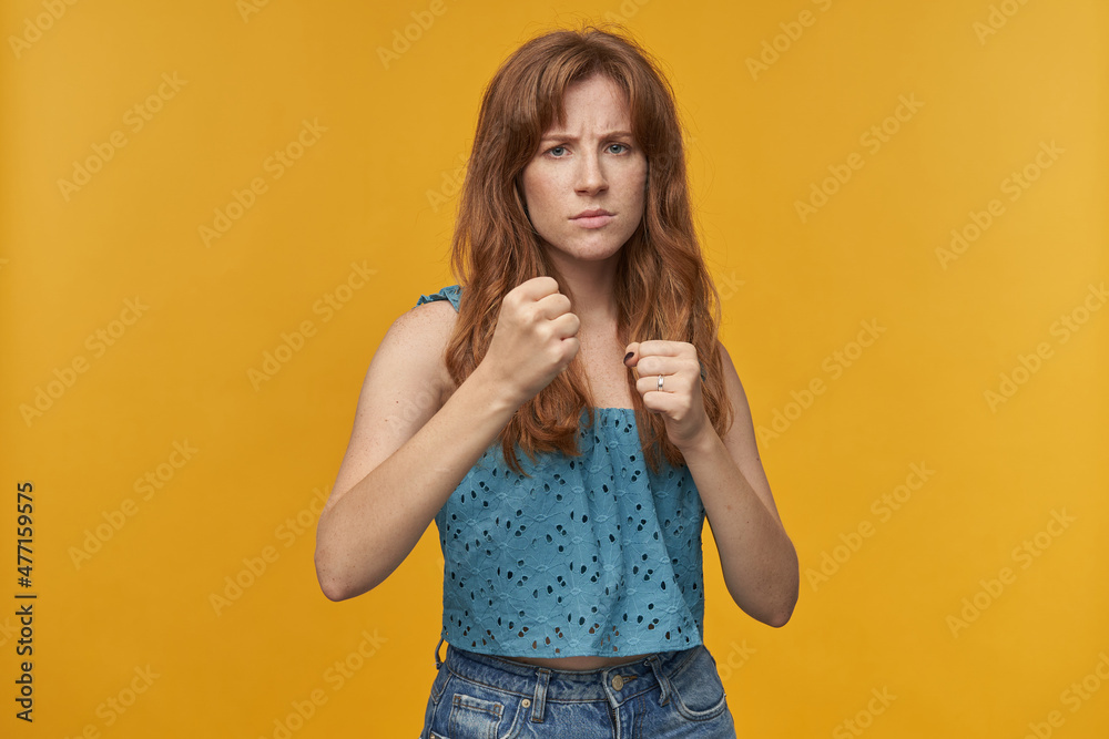 Indoor portrait of young redhead female with wavy long hair ready to fight for women rights, standing in boxing position isolated over orange background Indoor portrait of young redhead female with wavy long hair ready to fight for women rights, standing in boxing position isolated over orange background