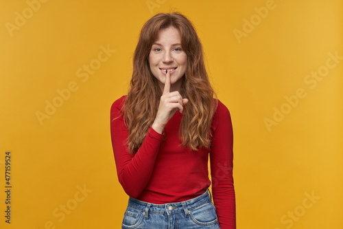 Indoor portrait of young redhead female with wavy long hair looks directly into camera, smiling and shows silence gesture, isolated over orange background