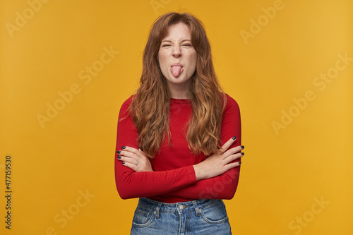 Indoor portrait of young redhead female with wavy long hair keeps her hands crossed, playfully shows her tongue into camera. isolated over orange background