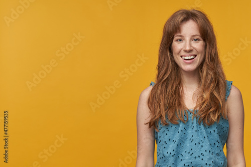 Indoor portrait of young redhead female with wavy long hair looks into camera and smiles broadly with copy space, isolated over orange background