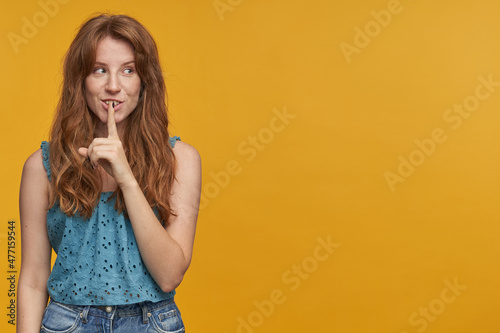 Indoor portrait of young redhead female with wavy long hair looks directly into camera, smiling and shows silence gesture, isolated over orange background