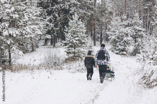 Rear view of father and little son walking in winter snowy pine forest together. Young man tourist in yellow jacket and teenage boy walking in snowy spruce park. Wintertime activity outdoors.