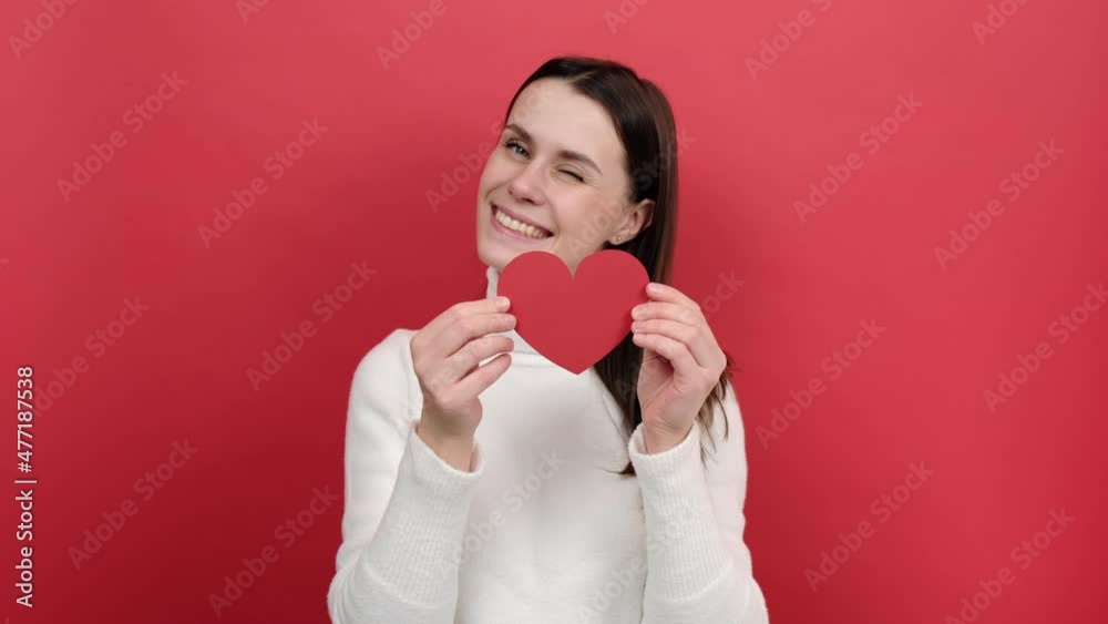Portrait of happy romantic young brunette female 20s holding heart love symbol, sending air kissing, flirting, wears white knitted sweater, posing isolated over red color background wall in studio Portrait of happy romantic young brunette female 20s holding heart love symbol, sending air kissing, flirting, wears white knitted sweater, posing isolated over red color background wall in studio