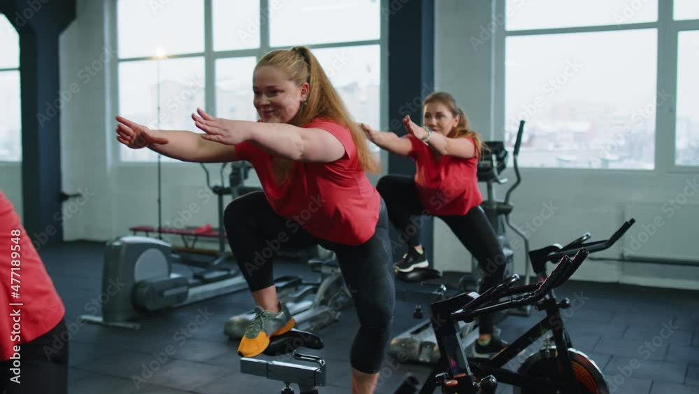 Group of smiling friends at gym exercising on stationary bike. Happy cheerful athletes training on exercise bike. Young women working out at class in gym. Weight loss, fat burning. Modern active sport Group of smiling friends at gym exercising on stationary bike. Happy cheerful athletes training on exercise bike. Young women working out at class in gym. Weight loss, fat burning. Modern active sport