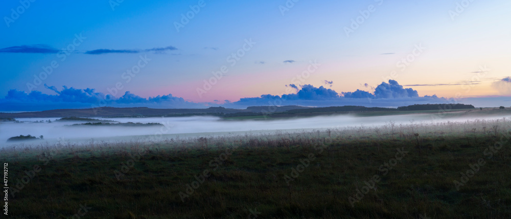 a blanket of ground fog rolling across meadows and woodland in the valley with a red, pink, orange and blue sunset sky, Wiltshire UK