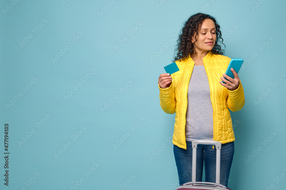 Young woman in yellow jacket stands against blue wall background, in front of her suitcase, holds mobile phone and plastic card with blank space for ad. Travel, booking, banking concept Young woman in yellow jacket stands against blue wall background, in front of her suitcase, holds mobile phone and plastic card with blank space for ad. Travel, booking, banking concept