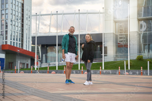 Full length shot of sportive middle aged man and woman in sportswear looking at camera while standing together outdoors after workout