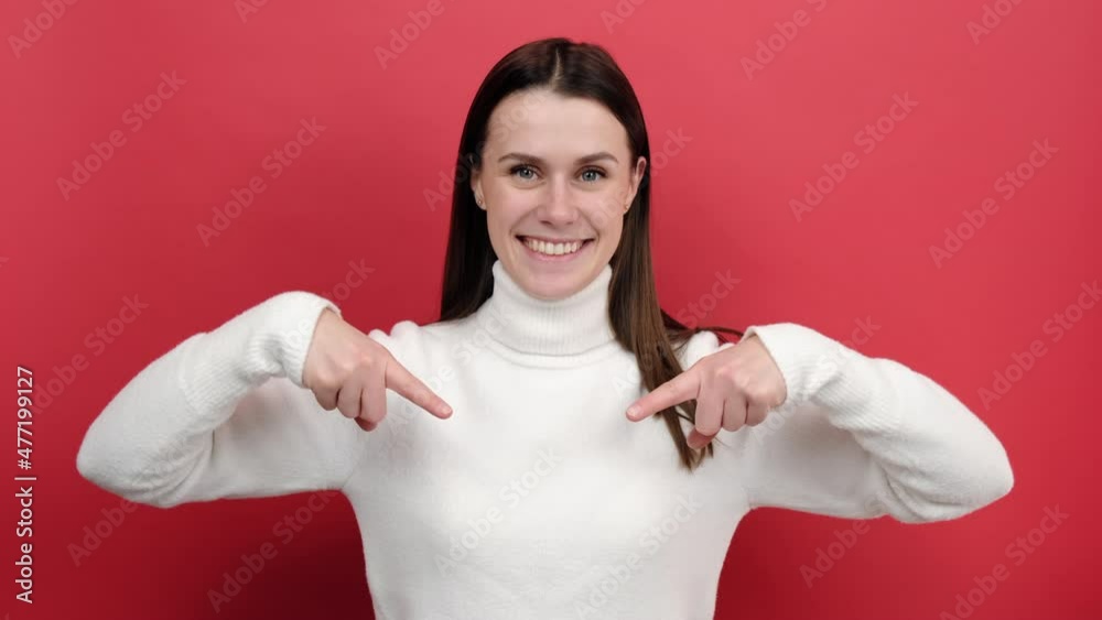 Portrait of happy young woman 20s pointing fingers down, asking to subscribe and like her post, recommending advertising area, wears knitted sweater, posing isolated over red color background studio