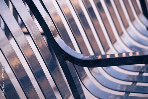 abstract view of a black public park bench reflecting sunset tones with focus on arm rest, repeating diagonal stripe pattern, and detailed metallic texture