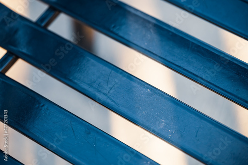 abstract closeup of a metallic public park bench seat creating diagonal stripes with blue tones against a light orange background