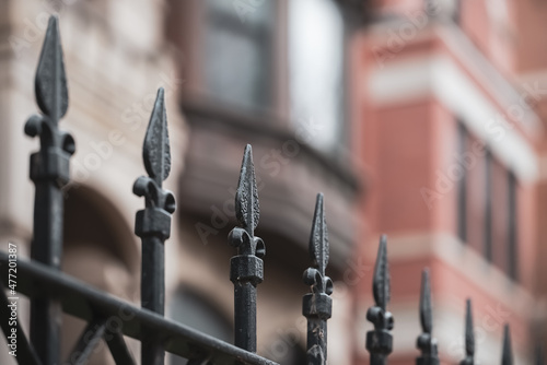 closeup of repeating ornate iron fence with selective focus on one spire, with blurred brown townhouses in the background