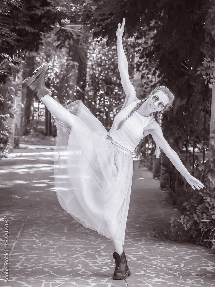 Naklejka premium A girl in a tutu and rough boots makes a leg swing with her arms spread wide on an alley in the park. Black and white photo. Blur focus