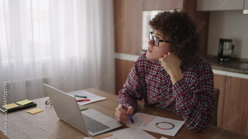 Brooding serious curly freelancer man sit at table in comfortable home office room work on laptop looks concentrated thinking over business issue solution, makes telecommute job