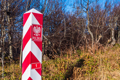 Polish, white and red border post. On the pole, there is a white eagle with a golden crown and the inscription Poland and number 1. In the background, the blue sky and trees, bushes and grass.