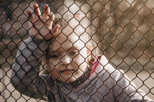 Little girl with a sad look behind a metal fence, social problems, raising children in orphanages