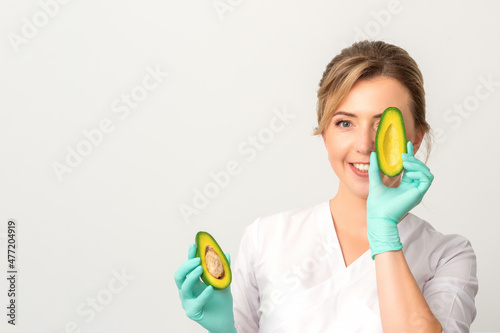 Portrait of young female nutritionist doctor with beautiful smile posing at camera hiding eye behind half avocado on white background, copy space. Benefits of proper nutrition