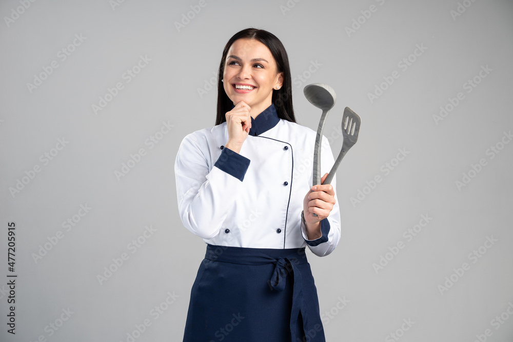 Portrait beautiful caucasian woman wearing uniform holding kitchen appliances and dreaming on grey isolated background. Occupation concept Portrait beautiful caucasian woman wearing uniform holding kitchen appliances and dreaming on grey isolated background. Occupation concept