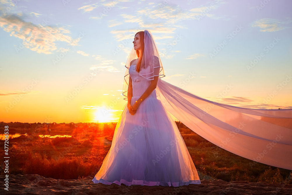 Beautiful bride in white dress and long fabric in the desert on sand on dunes and blue sky with yellow and red sunset
