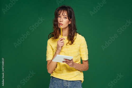 Pensive young girl with notebook decide problem, lost in thoughts looking aside on empty space on green background © Georgii