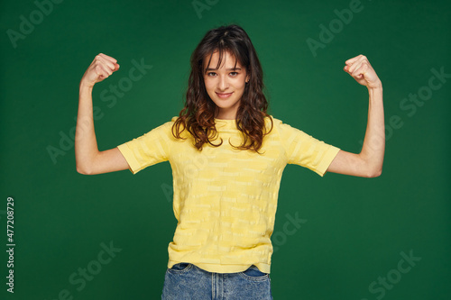 Pretty young girl show hands muscles, power, feel proud about achievements, keeps fit and healthy on green background © Georgii