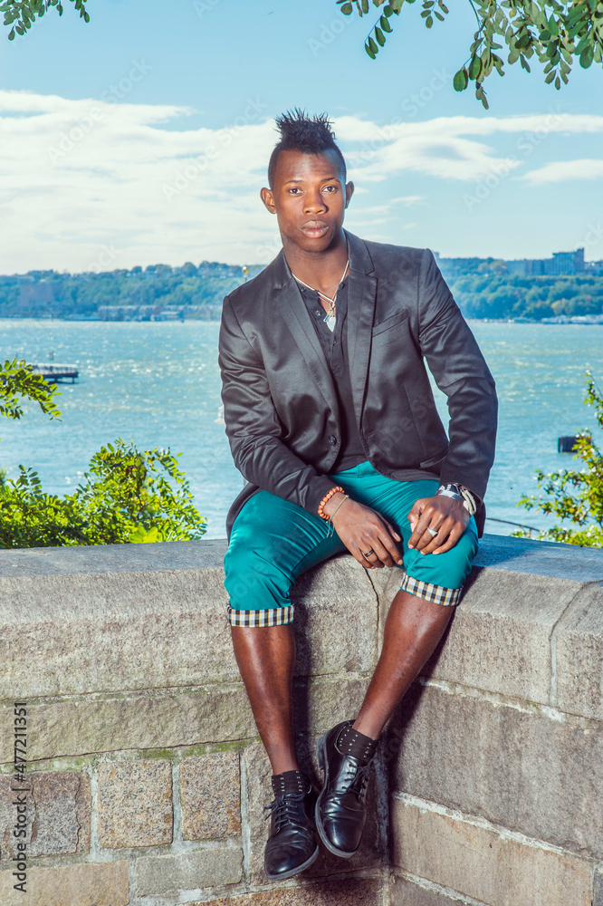 Man Relaxing Outside. Dressing in a black blazer green pants leather shoes a young black guy with mohawk hair is sitting on a rocky fence by a river relaxing. Urban Fashion. Photos