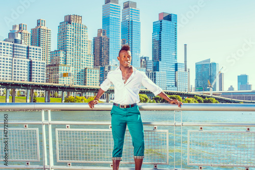 Urban Casual Fashion. Dressing in a white, long sleeve shirt, green pants, a young black guy with mohawk haircut is standing in the front of business district, confidently looking forward..