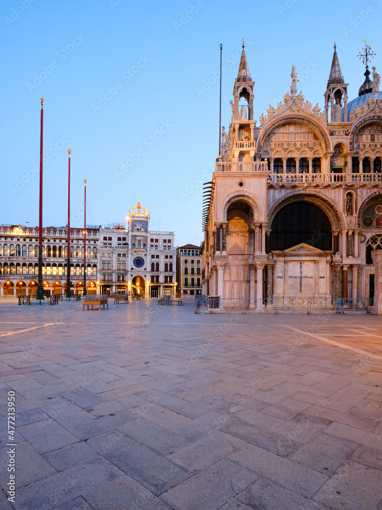 Basilica in San Marco square in Venice at dawn, on sunrise, illuminated with orange sunlight. Illuminated house with astronomical clock. Copy-space on stones of the square.