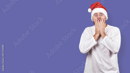 a young man in a Santa Claus hat covers his mouth with his hands in shock