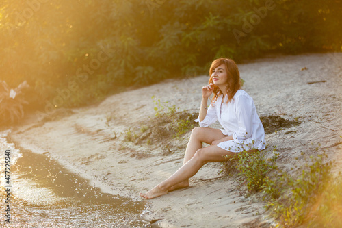 Portrait of young dreaming happy smiling woman weared white shirt and holding white hat walking near a lake or pond. Summer weekend or vacation story. Brown-haired girl with long hairs. Local tourism 