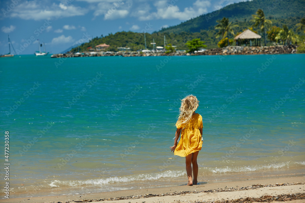 A young girl in a bright yellow dress admires the turquoise waters at the beach with palm trees