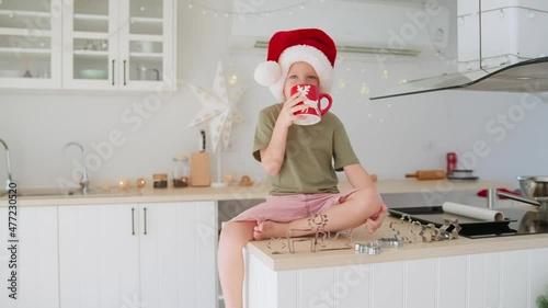 Little boy in Santa hat sitting on the kitchen table and drinking cocoa milk.