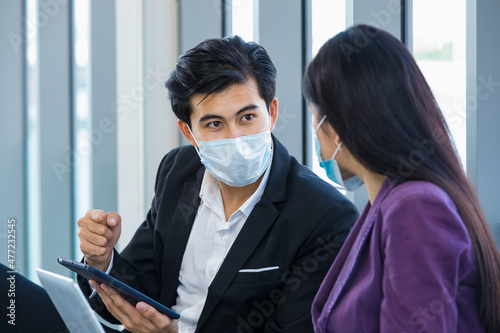 Millennial Asian male professional successful businessman manager in formal suit wearing safety face mask sit talking discussing via tablet and laptop computer with businesswoman colleague in office photo