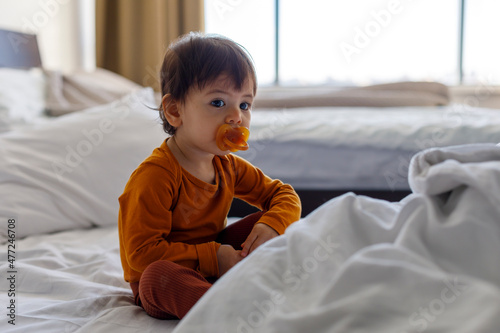 A one-year-old child with a pacifier in his mouth is sitting on the bed in the bedroom.