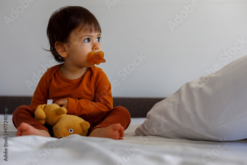 A one-year-old child with a pacifier in his mouth and a soft toy is sitting on the bed in the bedroom.