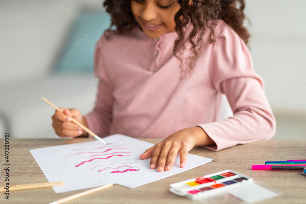 Inspired african american teen girl with brush and palette painting, enjoying her new hobby, sitting at desk at home Inspired african american teen girl with brush and palette painting, enjoying her new hobby, sitting at desk at home
