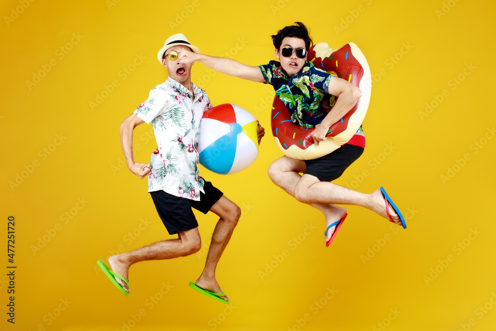 Two young Asian guys in summer casual with hat and sunglasses jumping up high with funny gesture with beach ball and ring float