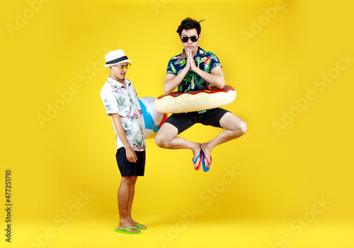 Young Asian guy in fancy summer dress, black glasses, and swimming float jumping up high and put hands to pay respect while happy friend with pool ball playing outdoor fun together